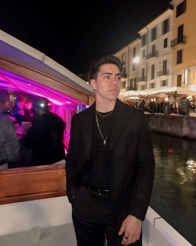 Matthew Stevens in a black suit beside a canal at night, with city lights behind