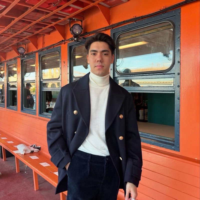 Matthew Stevens in a navy double-breasted coat and white roll-neck against a bright orange wall